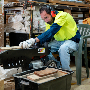 Worker in hi-vis and ear protection operating a table saw in a timber workshop.